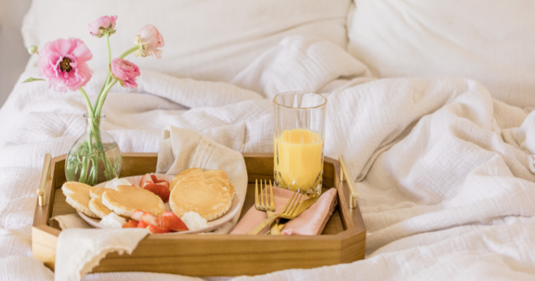 Light pink bedding background. A wood tray on the bedding containing a heart shaped pancake breakfast with orange juice and flowers on it.