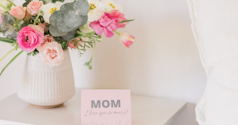 Light pink background. Small table with a vase of flowers and a card that says "Mom I love you so much!"